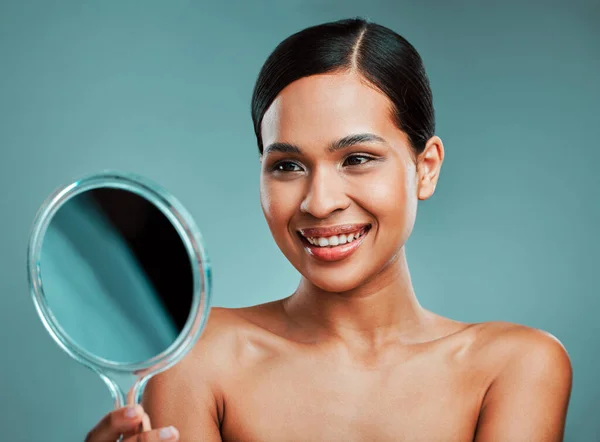 A beautiful young mixed race woman admiring herself in the mirror against a green studio background. Latin female smiling and looking at her reflection in a mirror.