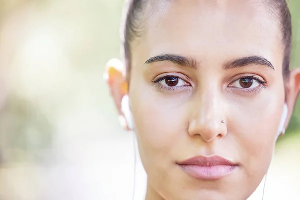 Close up face of sporty woman listening to music with earphones while standing outdoors. Portrait of beautiful hispanic woman with brown eyes and nose piercing.