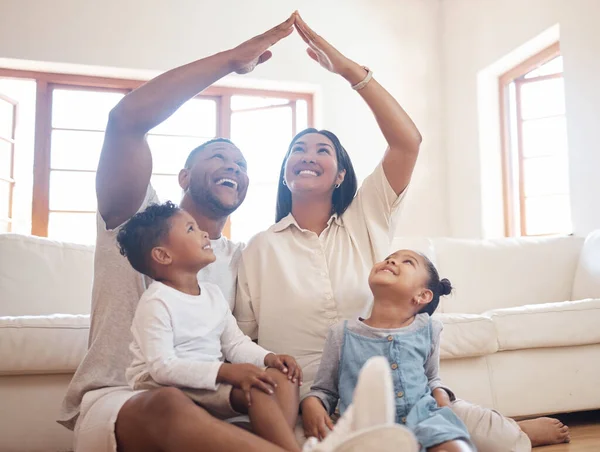 Stylish mixed race parents with two kids sitting on floor, mom and dad making roof figure with hands arms over heads of adorable son and daughter inside their new house.