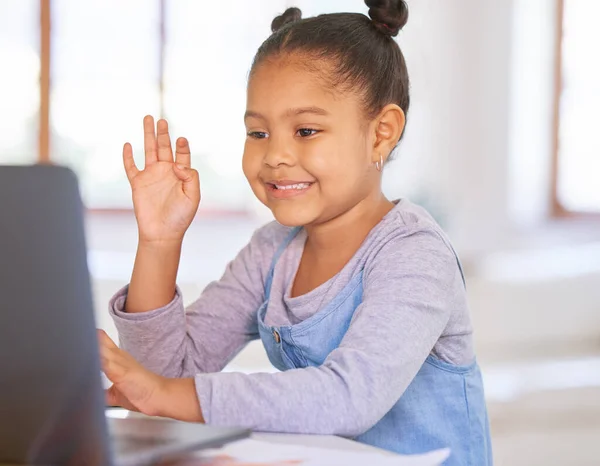 One happy mixed race preschool girl waving to teacher or tutor while having a video call on a laptop for distance learning at home. School kid attending online virtual education class for homeschool.