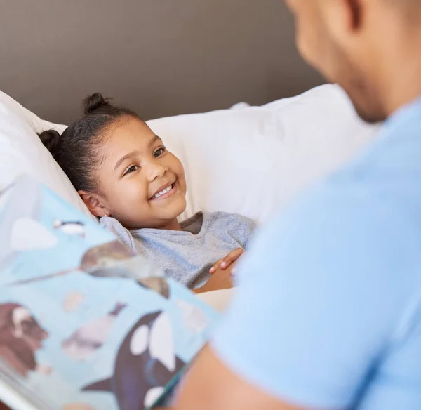 A cute little smiling mixed race girl laughing while lying in bed and enjoying story time. Young father reading his adorable african American daughter a bedtime story while she relax in bed at home.
