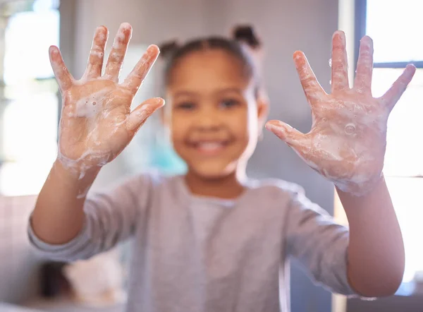 Little girl washing hands with water and soap in a bathroom. Happy kid showing soapy palms. Hand hygiene and virus protection. African American girl smiling while playing with foam from soapy hands.