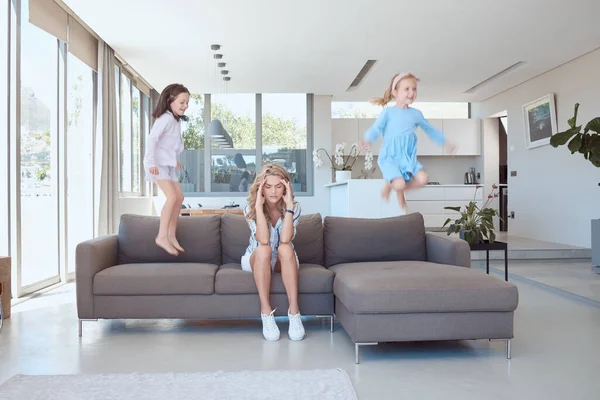 Fullbody caucasian woman sitting on a sofa at home and suffering from a headache while her hyperactive daughters jump around. Two adhd children annoying their mother by being naughty and misbehaving.