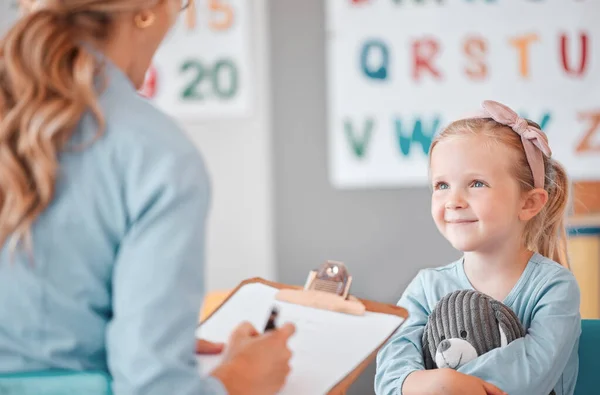 Young smiling caucasian child sitting with her psychologist in a clinic. Cute girl talking to a mental health professional. Using a clipboard to take notes on her patient. Dyslexia and anxiety.