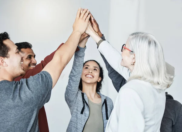Diverse group of five businesspeople smiling giving each other a high five in a meeting in an office at work. Happy women and men joining their hands in unity standing together while working.