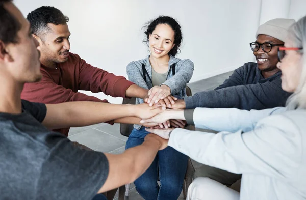 Diverse group of people sitting together in a circle and stacking their hands in the middle after therapy. Smiling support group celebrating after a successful session. Friends support mental health.