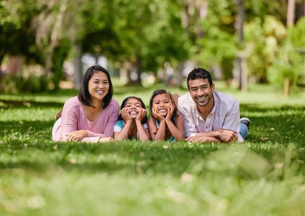 Young happy mixed race family relaxing and lying on grass together in a park. Loving parents spending time with their little daughters in nature. Joyful girls bonding with mom and dad outside.