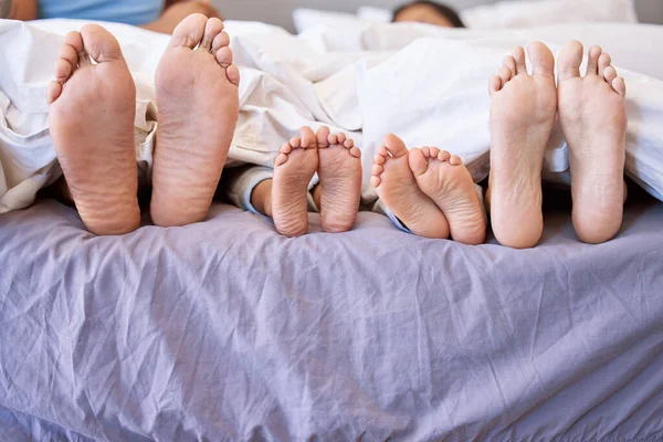 Feet of family lying in bed. Closeup of feet of parents and children side by side in bed. Family relaxing in bed together. Below bare feet of family in bed. Kids resting in bed with their parents