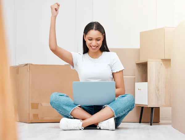 I got the deal. Full length shot of a young woman sitting in her new home and celebrating a success while using her laptop