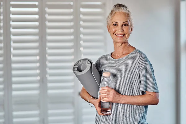 This is what I do every morning. Portrait of a cheerful mature woman holding a bottle of water and yoga mat ready to start her morning session of yoga inside of a studio