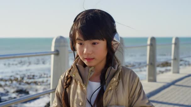 日本の休日を考えるビーチ遊歩道での旅行 女の子とヘッドフォン音楽 かわいいアジアの子供ストリーミングオーディオアプリで海思慮深く 熟考し 休暇について考える — ストック動画