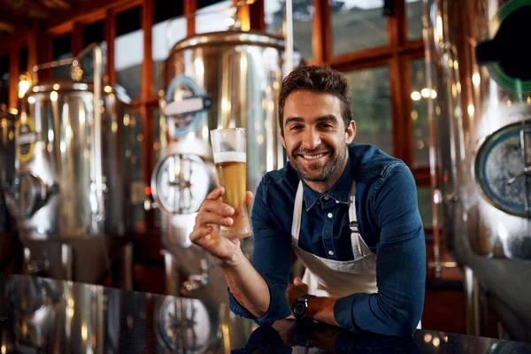 Who would like to have a taste. Portrait of a cheerful young business owner holding up a glass of beer that he just poured inside of a beer brewery during the day