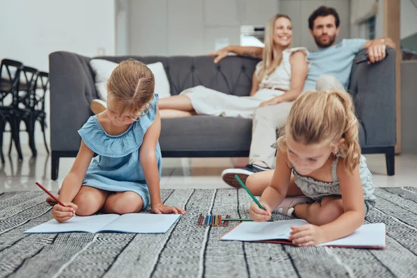 Living room, parents relaxing and children drawing in books on the floor in their modern home. Mother, father and girl kids sitting in the lounge together to rest, draw and bond in their house
