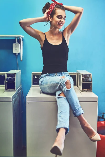 She even makes doing the laundry look sexy. an attractive young woman seated on a washing machine while holding her hair up and waiting for the washing to be done inside of a laundry room