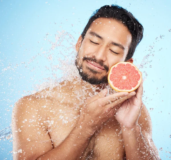 Face, water and grapefruit with a man model in studio on a blue background for hygiene or natural hydration. Skincare, beauty or fruit with a handsome young male wet from a water splash in the shower.