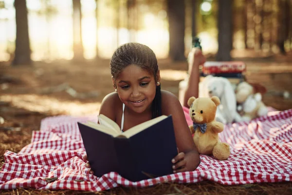 Shes a bookworm. a little girl reading a book with her toys in the woods