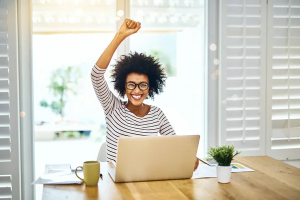 I am the real deal. Portrait of a cheerful and motivated young woman working on a laptop while putting her fist in the air at home