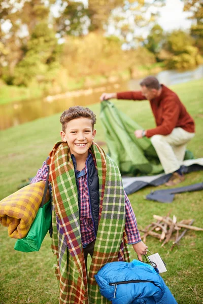 Im ready for anything on this camping trip. Portrait of a young boy holding camping gear with his father in the background
