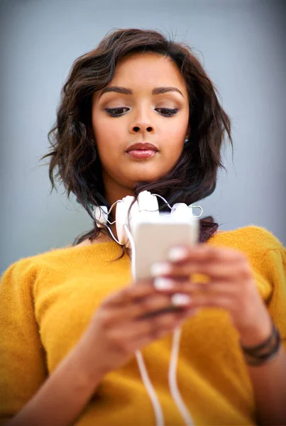 Waiting for a new song to download. Studio shot of a young woman with headphones around her neck using a mobile phone against a gray background