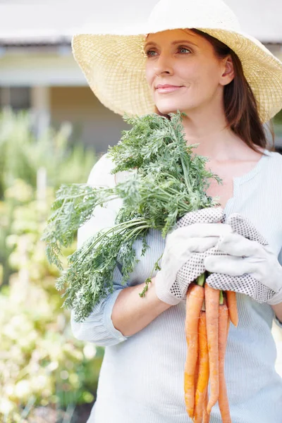 What is life without vegetables. A beautiful woman, wearing gardening gloves and a straw hat, holds a bunch of carrots while looking to the side