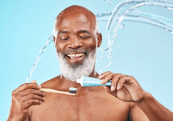 Water, splash and man with dental care in a studio for mouth health and wellness. Toothpaste, toothbrush and elderly African guy brushing his teeth for fresh oral hygiene isolated by blue background