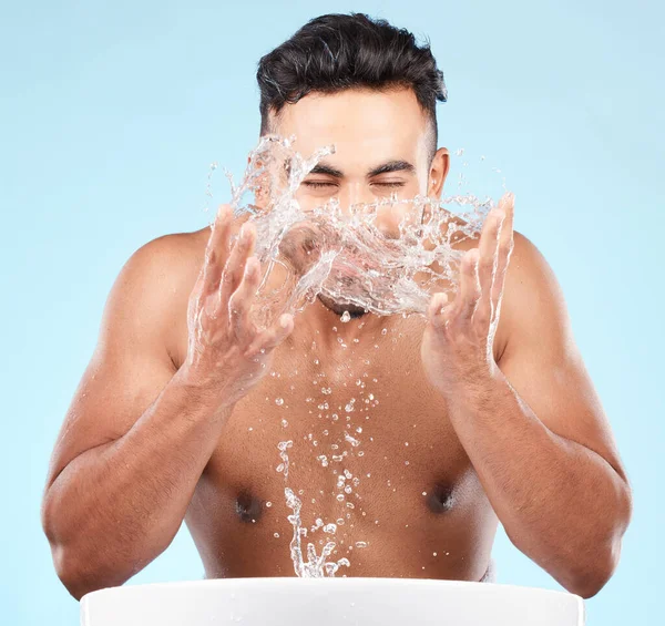 Face, water splash and skincare of man cleaning in studio isolated on a blue background. Hygiene, water drops and male model washing, bathing or grooming for healthy skin, facial wellness or beauty