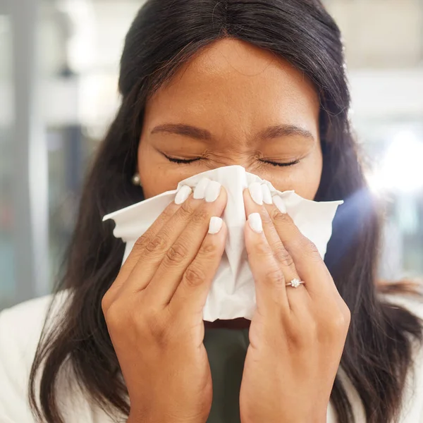 Woman, sneeze and tissue for flu, covid and safety in workplace with hands on face for health by blurred background. Corporate black woman, office and sick with toilet paper, nose and covid 19 virus.