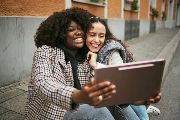City, friends and women take selfie on tablet while sitting on sidewalk laughing and happy together. Photo, video call and black woman with girl friend with urban fun and social media profile picture.