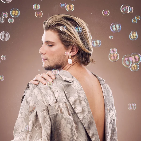 Beauty, lgbtq fashion and man with bubbles in studio on brown background for cosmetics, style and modern outfit. Creative art, luxury design and non binary model with soap bubbles, jewelry and makeup.