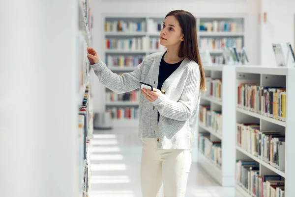 Library, book choice and student with phone in university, college or school. Learning, education scholarship or woman holding mobile looking for books by bookshelf for reading, knowledge or studying.