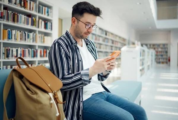 Library, university and man with phone on sofa for education, research and checking social media. Networking, knowledge and male student on smartphone, mobile app and website in college bookstore.