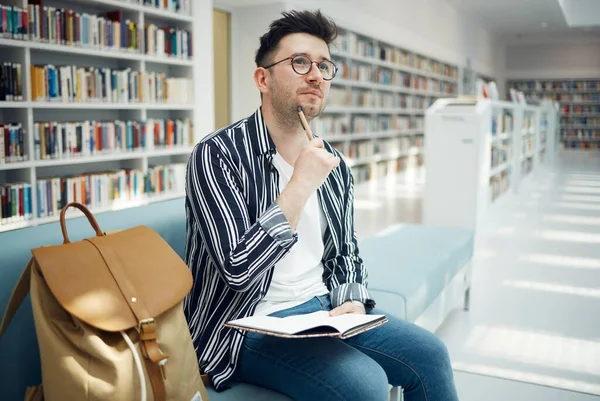University, student and thinking of idea in library for educational task with focus and notebook. Learning, knowledge and education of smart college man busy with assignment note ideas
