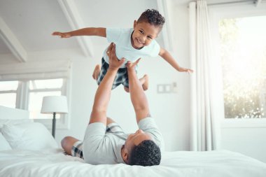 Cute little mixed race boy lying in fathers arms while looking at the camera and smiling. Loving family with father lifting happy little child son playing plane and having fun on bed. clipart