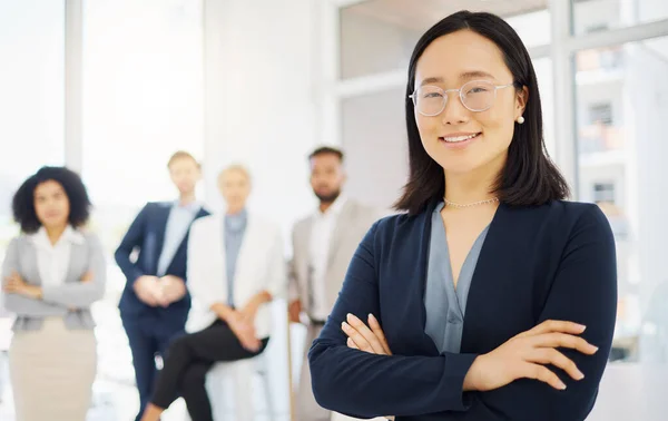 Portrait of a confident young asian businesswoman wearing glasses and standing with her arms crossed in an office with her colleagues in the background. Ambitious entrepreneur and determined leader r.