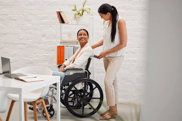 Young mixed race businesswoman helping a colleague in a wheelchair to her office at work. Hispanic businessperson with a disability being helped to her desk at work.