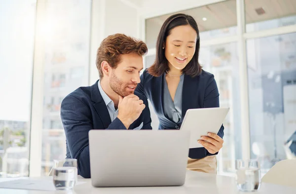 Two happy young diverse colleagues working together on a digital tablet in an office. Confident asian businesswoman showing smiling caucasian businessman corporate plans and ideas on device. Secretar.