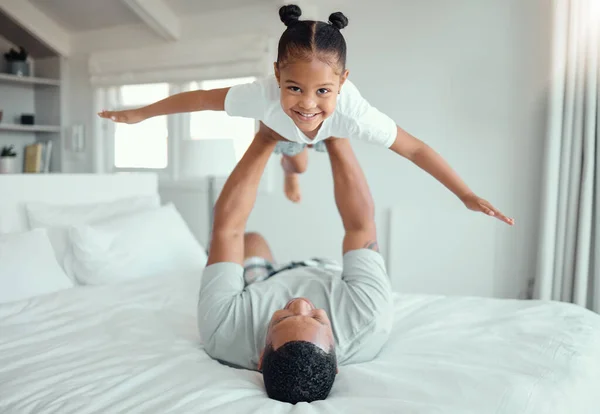 Happy little mixed race girl lying in fathers arms while looking at the camera and smiling. Loving family with father lifting cute little child daughter playing plane and having fun on bed.
