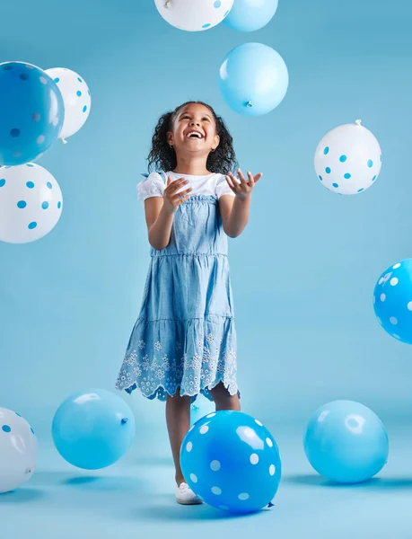 Adorable little girl looking up playing and having fun with blue and white balloons in celebration of her birthday against a blue studio background.