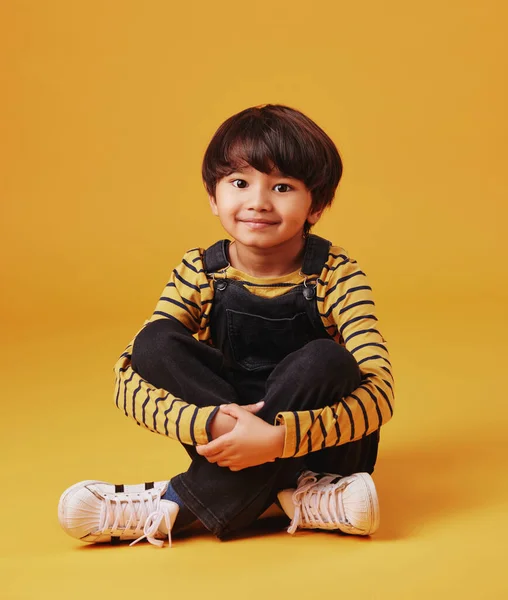 A cute little asian boy sitting on the floor with casual clothes while laughing and crossing his legs against an orange copyspace background. Adorable happy little boy safe and alone.