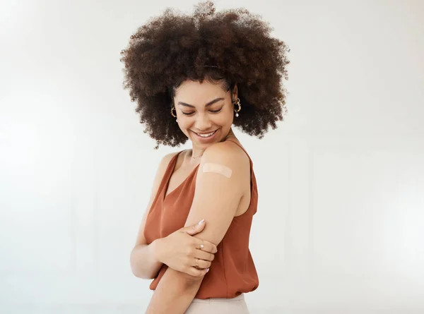 Young hispanic mixed race woman wearing a bandaid on her arm standing against a white studio background. Happy woman with a curly afro looking at a plaster on her arm against a white background.