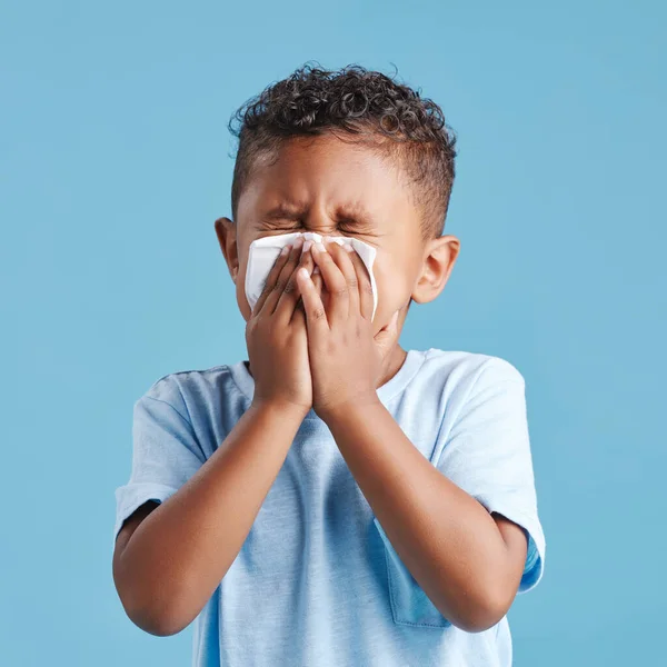 Little hispanic boy blowing his nose with tissue against blue studio background. Child feeling sick with flu virus or infection and suffering with snotty nose.