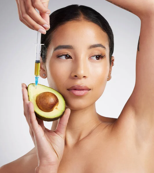 A gorgeous mixed race woman holding an avocado and syringe. Hispanic model promoting the skin benefits of avocado extract against a grey copyspace background.