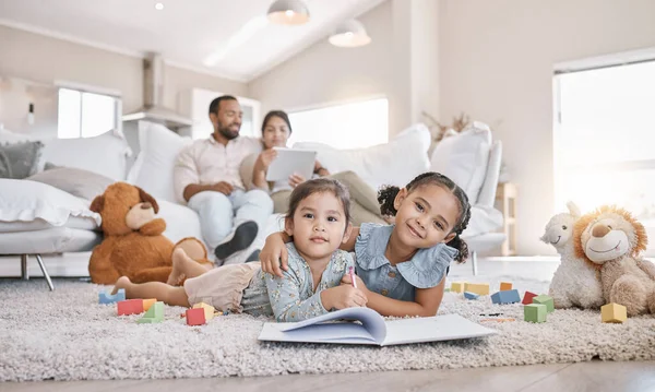 Little two girls drawing with colouring pencils lying on living room floor with their parents relaxing on couch. Little children sisters siblings colouring in during family time at home.