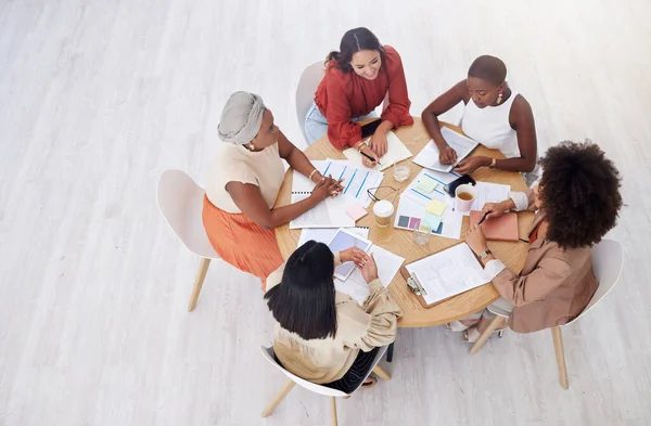 Above view of diverse group of business women having a brainstorm meeting in office with copyspace. Confident professional team sitting together and using paperwork while planning a marketing strateg.