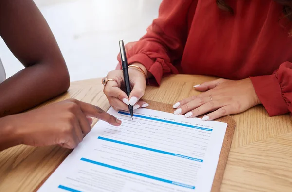 Closeup of two unknown ethnic business woman sitting and signing office contract. African american professional using hand gesture to point on paperwork. Mixed race colleague agreeing to document dea.