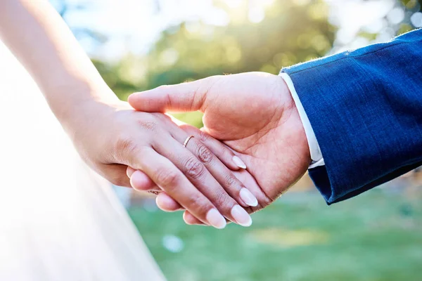Close up hands of newlywed couple standing outside on a sunny day. Groom holding his brides hand with wedding band.
