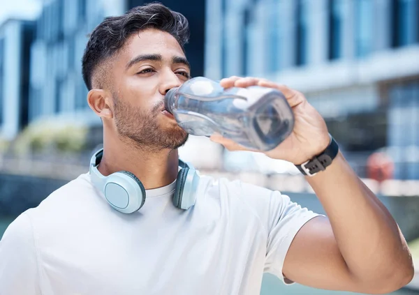 Young male athlete taking a break while drinking water from a bottle and wearing his headphones around his neck while out for a run and exercising outdoors during the day.