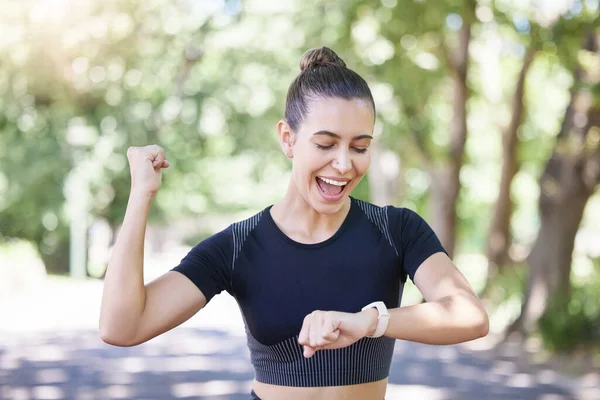 Cheerful young sporty female athlete celebrating while looking at smart watch. Hispanic sportswoman making winner gesture with clenched fist while tracking her progress while training outdoors.