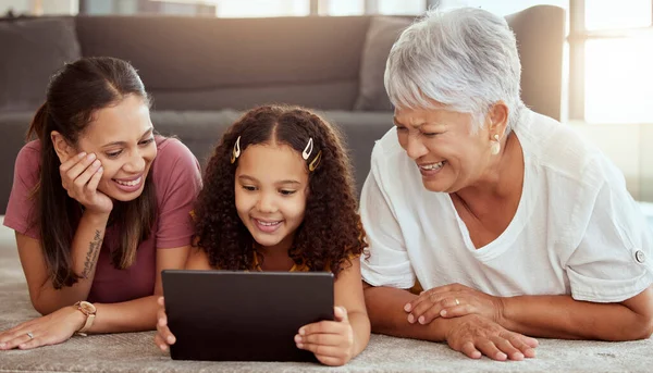 Adorable little mixed race child bonding with single mother and grandmother and using digital tablet at home. Parent and senior woman lying on living room floor together with kid and using technology.