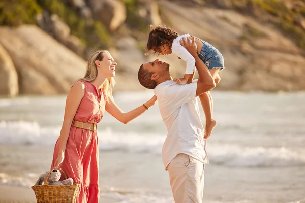 Full length of a happy mixed race family enjoying family time together at the beach. Loving father lifting his daughter in the air and having fun by the sea while her mother watch. Young couple enjoy.